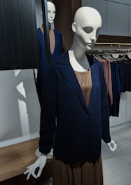 A mannequin is dressed in a formal outfit, consisting of a navy blue blazer over a brown dress. It is placed in a retail setting with another mannequin in the background, displaying a similar style. The environment includes clothing racks with garments in subdued colors, wood accents, and a clean, minimal aesthetic.