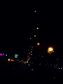 A night scene featuring a string of illuminated lanterns in various colors, beautifully lighting up the dark sky. There is a neon sign in the background, suggestive of an Indian restaurant, and additional small lights line what appears to be a street or pathway.