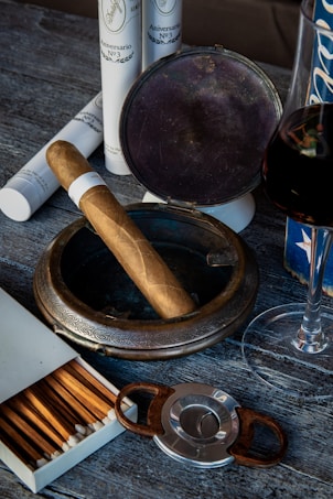 A rustic setting features a wooden surface with a silver, ornate ashtray holding a cigar. Nearby, a cigar cutter and an open box of wooden matches are visible. Several white, cylindrical cigar tubes are arranged in the background. A partially visible glass of red wine sits to the right, adding a touch of elegance.