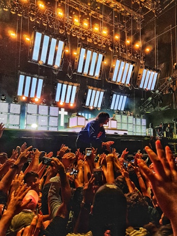 An energetic concert scene with a performer interacting with an enthusiastic crowd, hands reaching up towards the stage. The stage is illuminated with bright lights and large screens displaying patterns, creating a dynamic and electric atmosphere.