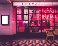 The image depicts the entrance of a cinema with illuminated neon signs. A person is seated at a table outside while another is visible inside, ascending the red stairs. The scene is bathed in pink and purple lighting, creating a vibrant and retro atmosphere.