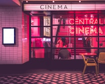 The image depicts the entrance of a cinema with illuminated neon signs. A person is seated at a table outside while another is visible inside, ascending the red stairs. The scene is bathed in pink and purple lighting, creating a vibrant and retro atmosphere.