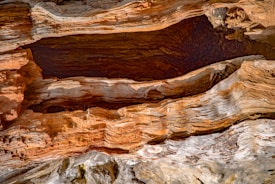 The image features the intricate texture of weathered wood, showcasing layers of rich brown and grey tones. The surface appears rough and uneven with distinct lines and grooves running through the wood, highlighting natural aging and weathering processes.