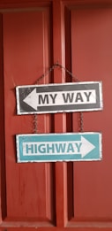 Two rectangular signboards are hanging on a chain against a red door. The top signboard has a white background with a black border and features a left-pointing arrow with the text 'MY WAY' in bold black letters. The bottom signboard has a blue background with a white border and displays a right-pointing arrow with the text 'HIGHWAY' in bold white letters.