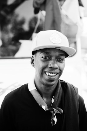A black and white portrait of a young person wearing a baseball cap and a scarf. They are smiling widely, exuding a sense of friendliness and approachability. The background is blurry, suggesting an outdoor setting with some structures or flags visible.