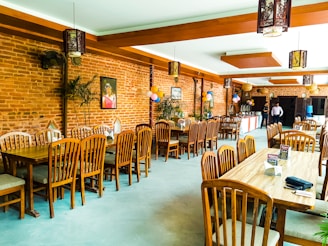 A restaurant interior featuring wooden tables and chairs arranged neatly. The walls are made of exposed brick, adorned with a few paintings and decorative elements. Balloons are strategically placed, suggesting a celebratory event. Warm lighting fixtures hang from the ceiling, enhancing the cozy ambiance. A buffet table is set up in the background with two people standing nearby.