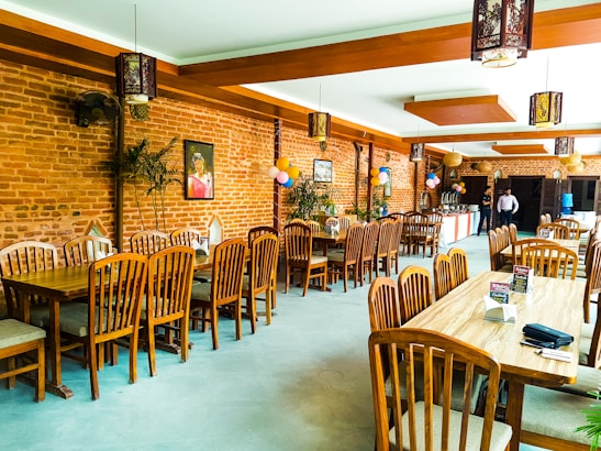 A restaurant interior featuring wooden tables and chairs arranged neatly. The walls are made of exposed brick, adorned with a few paintings and decorative elements. Balloons are strategically placed, suggesting a celebratory event. Warm lighting fixtures hang from the ceiling, enhancing the cozy ambiance. A buffet table is set up in the background with two people standing nearby.