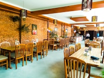 A restaurant interior featuring wooden tables and chairs arranged neatly. The walls are made of exposed brick, adorned with a few paintings and decorative elements. Balloons are strategically placed, suggesting a celebratory event. Warm lighting fixtures hang from the ceiling, enhancing the cozy ambiance. A buffet table is set up in the background with two people standing nearby.