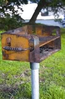 A rusty metal barbecue grill stands outdoors on a metal pole in a grassy area. The grill is weathered, showing signs of wear and oxidation with a spiraled handle. In the background, there are blurred trees and a hint of a road.