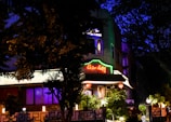 A building is illuminated at night, featuring a bright neon sign that reads 'China Valley'. The structure is surrounded by trees and foliage, with lamps enhancing the warm ambiance. Decorative red lanterns hang in front of the entrance, contributing to an inviting atmosphere.