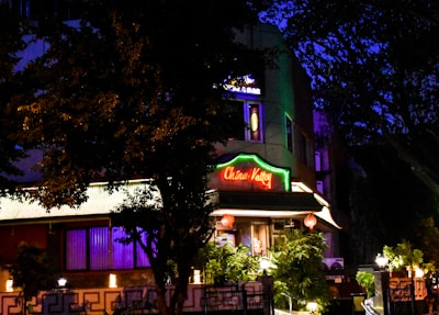 A building is illuminated at night, featuring a bright neon sign that reads 'China Valley'. The structure is surrounded by trees and foliage, with lamps enhancing the warm ambiance. Decorative red lanterns hang in front of the entrance, contributing to an inviting atmosphere.