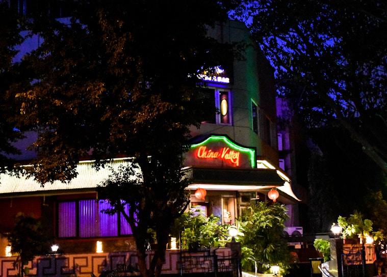 A building is illuminated at night, featuring a bright neon sign that reads 'China Valley'. The structure is surrounded by trees and foliage, with lamps enhancing the warm ambiance. Decorative red lanterns hang in front of the entrance, contributing to an inviting atmosphere.