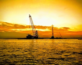 A large construction crane stands against a vibrant orange and yellow sunset. The silhouetted crane is positioned on a body of water, likely in an industrial or harbor setting. Ripples on the water surface reflect the warm hues of the sky, complementing the overall serene yet industrious scene.