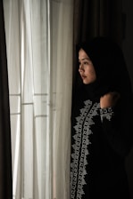 A woman wearing a dark, embroidered hijab stands thoughtfully by a window with sheer white curtains. Soft light filters through the fabric, casting a gentle illumination on her face.