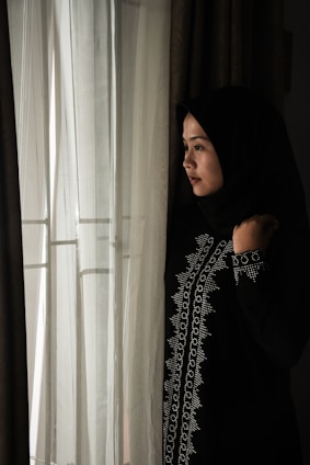 A woman wearing a dark, embroidered hijab stands thoughtfully by a window with sheer white curtains. Soft light filters through the fabric, casting a gentle illumination on her face.