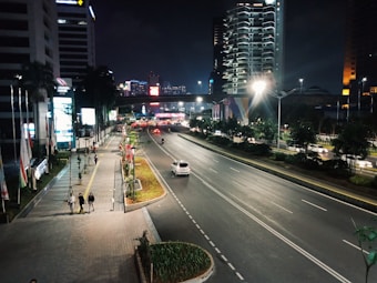 A nighttime cityscape with modern tall buildings illuminated against a dark sky. A wide road with minimal traffic stretches out, flanked by trees and pedestrian pathways. Bright streetlights and various colored lights from billboards and buildings create a vibrant urban scene.