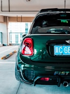 A dark green, compact car is parked in an indoor garage. The focus is on the car's rear, showing a shiny surface, taillights, and a close-up of the license plate with foreign characters. The environment is modern and well-lit, with concrete flooring and minimal barriers.