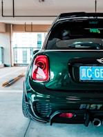 A dark green, compact car is parked in an indoor garage. The focus is on the car's rear, showing a shiny surface, taillights, and a close-up of the license plate with foreign characters. The environment is modern and well-lit, with concrete flooring and minimal barriers.