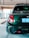 A dark green, compact car is parked in an indoor garage. The focus is on the car's rear, showing a shiny surface, taillights, and a close-up of the license plate with foreign characters. The environment is modern and well-lit, with concrete flooring and minimal barriers.