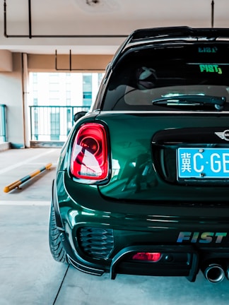 A dark green, compact car is parked in an indoor garage. The focus is on the car's rear, showing a shiny surface, taillights, and a close-up of the license plate with foreign characters. The environment is modern and well-lit, with concrete flooring and minimal barriers.
