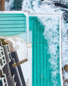 An aerial view of an ocean-side swimming pool where waves crash over the edge, filling it with seawater. The pool has distinct lap lanes, and a solitary swimmer is visible. Surrounding the pool, there are terraces and walkways, with a building structure on one side.