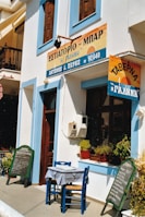 The image depicts the exterior of a quaint restaurant or bar with Greek signage, situated on a sunlit street. A small table and chairs are placed outside, featuring a checkered tablecloth. The facade is adorned with blue and white accents, while potted plants add a touch of greenery by the window. A pair of chalkboards lists menu items in both English and Greek.