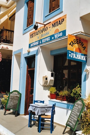 The image depicts the exterior of a quaint restaurant or bar with Greek signage, situated on a sunlit street. A small table and chairs are placed outside, featuring a checkered tablecloth. The facade is adorned with blue and white accents, while potted plants add a touch of greenery by the window. A pair of chalkboards lists menu items in both English and Greek.
