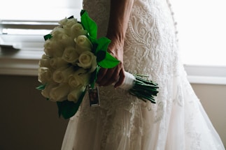 A person holding a bouquet of white roses with green leaves. The person is wearing an intricately designed lace wedding dress, with the details of the fabric visible. Natural light filters in from the background, creating a soft, romantic atmosphere.