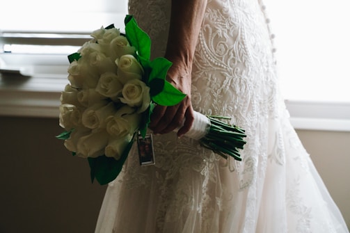 A person holding a bouquet of white roses with green leaves. The person is wearing an intricately designed lace wedding dress, with the details of the fabric visible. Natural light filters in from the background, creating a soft, romantic atmosphere.