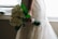 A person holding a bouquet of white roses with green leaves. The person is wearing an intricately designed lace wedding dress, with the details of the fabric visible. Natural light filters in from the background, creating a soft, romantic atmosphere.