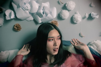 A person with long dark hair appears surrounded by an artistic arrangement of large paper flowers attached to a wall. The individual wears bold makeup with red eye shadow and is dressed in a sheer, colorful outfit. The background features white and gold-colored hanging decorations, creating a whimsical atmosphere.
