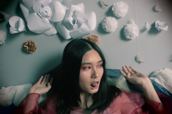 A person with long dark hair appears surrounded by an artistic arrangement of large paper flowers attached to a wall. The individual wears bold makeup with red eye shadow and is dressed in a sheer, colorful outfit. The background features white and gold-colored hanging decorations, creating a whimsical atmosphere.