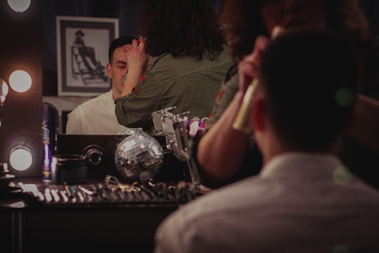 A person with short hair is seated in front of a vanity mirror, having makeup applied by another person. The mirror is surrounded by lights, and there are various cosmetic products and tools scattered on the tabletop. A silver disco ball and framed photograph are visible on the table.