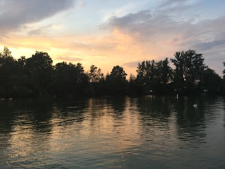 A serene lake reflecting the colorful sky at dusk, surrounded by dense forest.