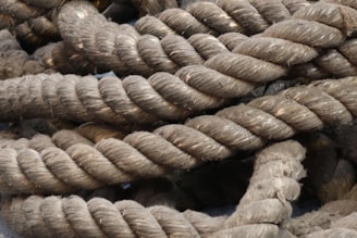 Thick, coiled ropes with a rough texture and a fibrous appearance are piled together. The ropes are predominantly a natural brown color with visible wear and strands unraveling slightly in some areas.