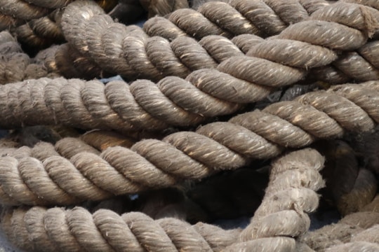 Thick, coiled ropes with a rough texture and a fibrous appearance are piled together. The ropes are predominantly a natural brown color with visible wear and strands unraveling slightly in some areas.