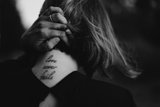 A close-up view of the back of a person's neck, adorned with a tattoo that reads 'love never fails.' A hand with a ring is gently holding the person's hair. The scene is captured in black and white, creating a serene and intimate atmosphere.