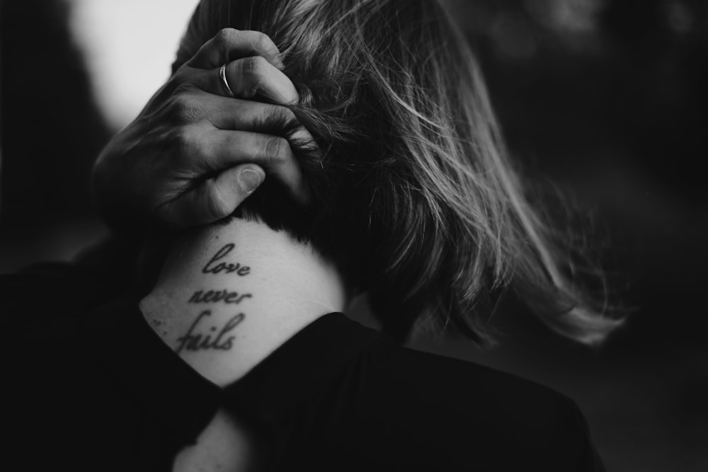 grayscale photo of woman holding her neck while showing Love never Fails tattoo at back