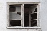A weathered and dilapidated window with shattered glass panes against a textured, worn wall.
