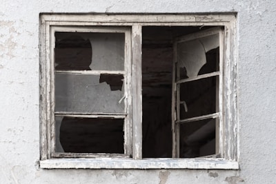 A weathered and dilapidated window with shattered glass panes against a textured, worn wall.