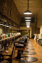 A long, narrow barbershop interior featuring a row of vintage-style barber chairs and mirrored stations along one wall. The ceiling is adorned with a series of pendant lights, providing warm illumination. The walls are rustic with exposed brick, adding a touch of old-world charm. Various hair care products are neatly arranged on the countertop.