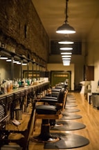 A long, narrow barbershop interior featuring a row of vintage-style barber chairs and mirrored stations along one wall. The ceiling is adorned with a series of pendant lights, providing warm illumination. The walls are rustic with exposed brick, adding a touch of old-world charm. Various hair care products are neatly arranged on the countertop.