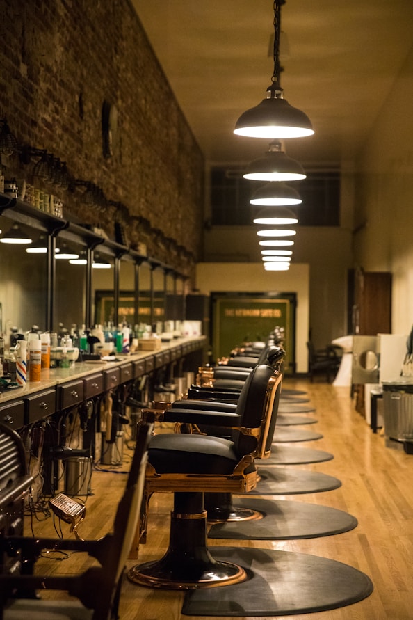 A long, narrow barbershop interior featuring a row of vintage-style barber chairs and mirrored stations along one wall. The ceiling is adorned with a series of pendant lights, providing warm illumination. The walls are rustic with exposed brick, adding a touch of old-world charm. Various hair care products are neatly arranged on the countertop.