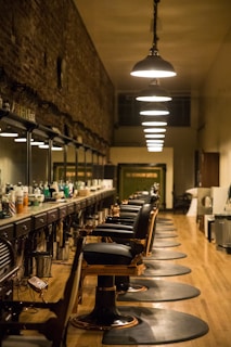 A long, narrow barbershop interior featuring a row of vintage-style barber chairs and mirrored stations along one wall. The ceiling is adorned with a series of pendant lights, providing warm illumination. The walls are rustic with exposed brick, adding a touch of old-world charm. Various hair care products are neatly arranged on the countertop.