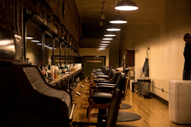 A well-lit, clean, and organized barbershop with multiple empty barber chairs aligned neatly along a counter. The counter is filled with various hair styling products and tools. The floor is made of polished wood, and overhead lights hang from the ceiling in a straight line providing warm illumination.