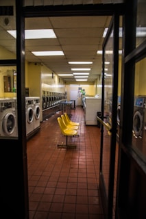 A long corridor of a laundromat with rows of washing machines on either side. The floor is made of reddish-brown tiles, and the ceiling is lined with fluorescent lights. In the center, there are several bright yellow plastic chairs facing each other.