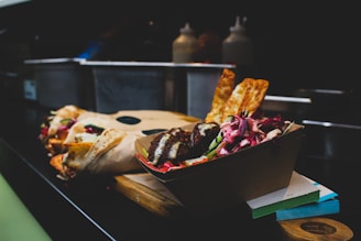 A selection of food items arranged on a wooden board, including a box filled with grilled meat, vibrant salad with purple cabbage, and crispy flatbread pieces. The background is a dimly lit kitchen setting with various containers slightly visible.