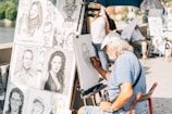 An elderly artist with a beard and cap is sketching a caricature at an outdoor setting. Multiple caricature and portrait drawings are displayed on a board next to him, depicting exaggerated features of various individuals. The artist is focused on his work, sitting under a blue umbrella on a sunny day, with passersby in the background.