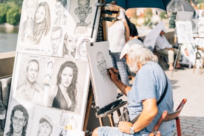 An elderly artist with a beard and cap is sketching a caricature at an outdoor setting. Multiple caricature and portrait drawings are displayed on a board next to him, depicting exaggerated features of various individuals. The artist is focused on his work, sitting under a blue umbrella on a sunny day, with passersby in the background.