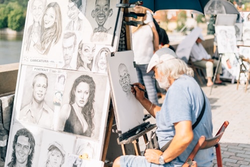 An elderly artist with a beard and cap is sketching a caricature at an outdoor setting. Multiple caricature and portrait drawings are displayed on a board next to him, depicting exaggerated features of various individuals. The artist is focused on his work, sitting under a blue umbrella on a sunny day, with passersby in the background.